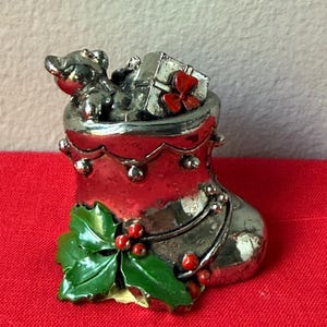 May include: A silver Christmas stocking figurine with a teddy bear and a gift box inside. The stocking has a red top and is decorated with holly leaves and berries. A red bow adorns the gift box.