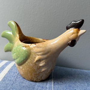 May include: A ceramic chicken-shaped pitcher with a tan body, green tail feathers, and a black comb. The pitcher has a small opening at the top and a spout for pouring. The design is rustic and charming, perfect for farmhouse decor.