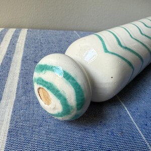 Op de afbeelding: Een witte keramische vaas met turquoise zigzagstrepen. De vaas heeft een afgeronde basis en een lange, cilindrische body. De vaas ligt op een blauw-wit gestreepte stof.