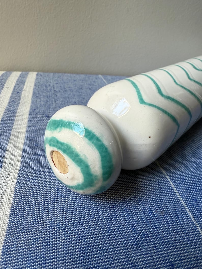 Op de afbeelding: Een witte keramische vaas met turquoise zigzagstrepen. De vaas heeft een afgeronde basis en een lange, cilindrische body. De vaas ligt op een blauw-wit gestreepte stof.