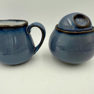 May include: A blue ceramic creamer and sugar bowl set. The creamer has a handle and spout, while the sugar bowl has a lid with a circular opening. Both pieces have a dark brown rim and a glossy finish, suitable for serving coffee or tea.