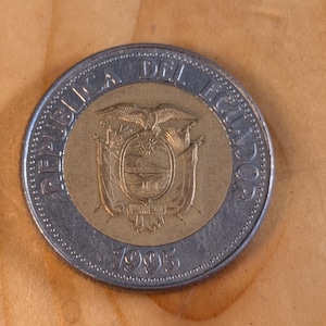 May include: A bi-metallic coin with a silver outer ring and a gold-coloured centre. The centre features a coat of arms design. The coin is dated 1995. The coin is on a wooden surface.