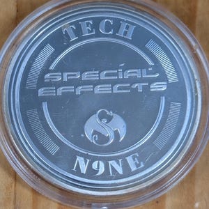 May include: A silver coin with the words "TECH SPECIAL EFFECTS" and "N9NE" in a clear plastic case. The coin has a detailed design and is set against a wooden background.