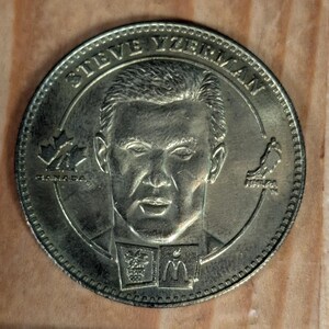 Peut inclure: Une pièce de monnaie commémorative dorée avec un portrait d'homme et le nom "STEVE YZERMAN" sur le bord. La pièce comporte divers symboles et logos, dont une feuille d'érable et un oiseau.