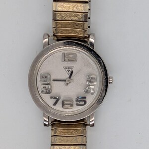 May include: A silver Guess wristwatch with a round face and a gold-toned, expandable strap. The watch face has large numerals and the Guess logo. The strap is made of interlocking metal links.