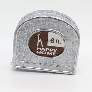 Puede incluir: Un flexómetro Happy Home plateado con una medida de 1,83 m. El flexómetro tiene una etiqueta ovalada marrón y blanca con el logotipo de Happy Home. El texto "ADD 2 INCHES" está impreso en el flexómetro.