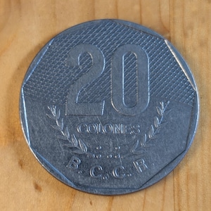 Puede incluir: Una moneda plateada con el número 20, la palabra "COLONES" y las letras "B.C.C.R." La moneda tiene una superficie texturizada y está sobre un fondo de madera. Una regla está en la parte inferior.