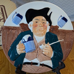 May include: A ceramic decorative plate depicting a man in a black hat, holding a mug and pipe. The plate has a brown and white colour scheme with blue accents. The man is dressed in a green coat and tan waistcoat.