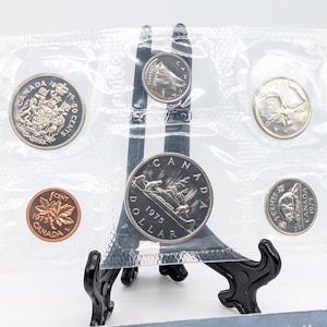 May include: A collection of Canadian coins from 1975, presented on a black stand. The coins are encased in a clear plastic sleeve. The set features a dollar coin, a 50-cent coin, a 25-cent coin, a 10-cent coin, and a 1-cent coin.