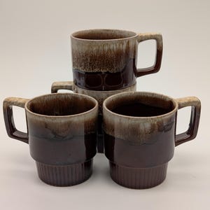 以下が含まれることがあります： 2色の釉薬が施された、4つの茶色のセラミックマグのスタック。マグは濃い茶色のベースと明るい茶色のトップで、正方形のハンドルが付いています。マグは積み重ねられています。