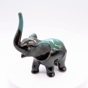 May include: A ceramic elephant figurine with a glossy finish. The elephant is primarily black with teal and brown accents on its trunk and back. The trunk is raised, and the figurine is on a white surface.