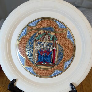May include: A cream-coloured decorative plate featuring a detailed, colourful illustration of two figures in blue robes and a church scene. The plate has a gold and blue border with an intricate pattern, displayed on a black stand.