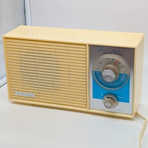 Könnte beinhalten: Ein cremefarbenes Vintage-Electra-Transistorradio. Das Radio hat einen Lautsprechergrill, ein blau-silbernes Zifferblatt und die Worte "Solid State Instant Sound" und "All Transistor". Das Radio hat ein cremefarbenes Netzkabel.