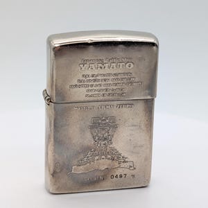 Może przedstawiać: Srebrny zapalniczka Zippo z wygrawerowanym tekstem i szczegółową ilustracją statku. Tekst brzmi "Japanese Battleship Yamato" i "World Army Zippo". Widoczny jest również numer seryjny 0467.