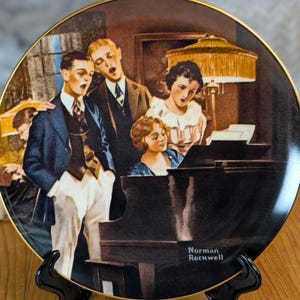 Puede incluir: Plato decorativo con una pintura de personas cantando alrededor de un piano. El plato tiene un borde dorado y se exhibe en un soporte negro. La pintura está firmada "Norman Rockwell".