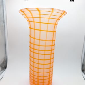 May include: A tall, orange glass vase with a grid pattern of orange lines. The vase has a flared top and a cylindrical body. The vase is sitting on a white, round platform.