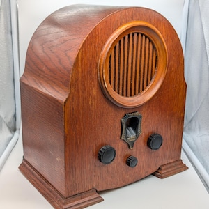 May include: A vintage wooden radio with a curved top and a circular speaker grill. The radio has a dark wood finish, black knobs, and a decorative metal plate. The radio is a classic piece of audio equipment.