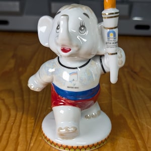 May include: A white ceramic elephant figurine holding a torch with an orange flame. The elephant wears a red, white, and blue outfit with the text "Yakutsk 2012" and Cyrillic text. The figurine stands on a round base.