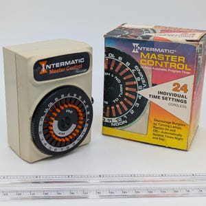 May include: An Intermatic Master Control timer with a black and orange dial, and its original box. The box features the Intermatic logo and text, "24 Individual Time Settings". The timer is cream-coloured.
