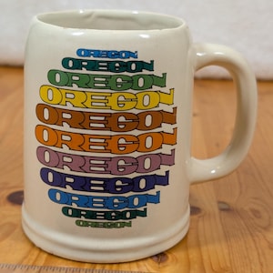 May include: A white ceramic mug with a handle. The mug has the word "OREGON" written on it in a rainbow of colors.