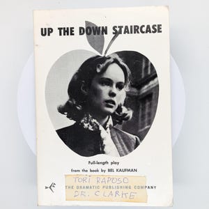 May include: Vintage paperback book titled "Up the Down Staircase." The cover features a black and white portrait of a woman within an apple-shaped design. Additional text includes the author's name, "Bel Kaufman," and "Tori Raposo."