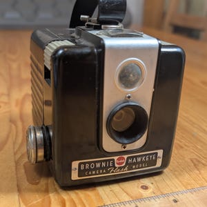 Puede incluir: Una cámara Kodak Brownie Hawkeye Flash modelo negro con un panel frontal plateado y un logotipo Kodak rojo. La cámara tiene un flash incorporado y un visor.