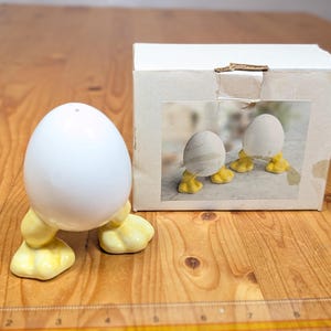 May include: Three white ceramic egg-shaped salt and pepper shakers with yellow feet. The shakers are on a wooden surface and are packaged in a white box with a photo of the shakers on the front.