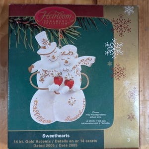 Peut inclure: Ornement de Noël en porcelaine blanche représentant deux bonhommes de neige tenant des cœurs rouges. L'ornement est décoré d'accents dorés et des mots "Sweethearts" et "2005".