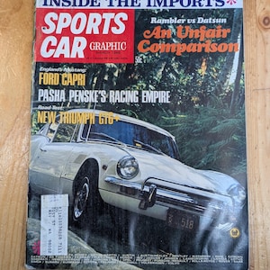 May include: A vintage issue of Sports Car Graphic magazine. The cover features a white car with the text "NEW TRIUMPH GT6+". Other text includes "INSIDE THE IMPORTS", "An Unfair Comparison", and "FORD CAPRI". The magazine is on a wooden surface.