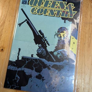 May include: A comic book titled "QUEENS & COUNTRY" in a protective plastic sleeve. The cover art features a soldier aiming a sniper rifle, with a blue and yellow color scheme. The comic is on a wooden surface.
