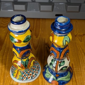 May include: Two decorative ceramic candle holders. Each is painted with bright colours including yellow, blue, orange and green. The bases have a patterned design. The top is a small, open cup shape with a blue rim.