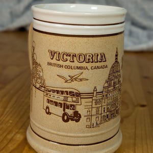 Op de afbeelding: Beige keramische bierpul met een witte rand en handvat. De pul is versierd met bruine illustraties van een dubbeldekkerbus, gebouwen en de tekst "VICTORIA British Columbia, CANADA".