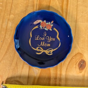 May include: A small, round, cobalt blue dish with a scalloped edge. The dish features a gold-colored decorative frame with the words "I Love You Mom" and two red roses. The dish measures approximately 3 inches in diameter.