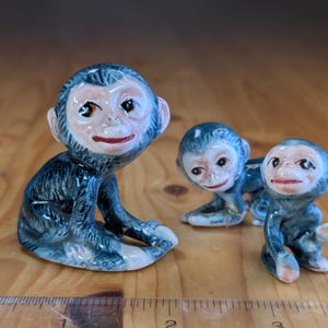 Peut inclure: Trois figurines de singes en céramique. Les singes ont une fourrure bleue, des visages bronzés et des lèvres rouges. Un singe est assis droit, tandis que les deux autres sont en position de ramper. Les figurines sont sur une surface en bois.
