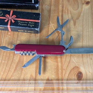 May include: A red multi-tool knife with various implements, including a blade, scissors, and a corkscrew. The knife is open, displaying its features. A box with the text "Superior Multi Purpose Knife" is visible.