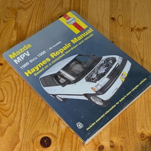 Puede incluir: Un manual de reparación Haynes para el Mazda MPV, modelos de 1989 a 1998. La portada muestra un coche blanco con el capó abierto, mostrando el motor. El libro está sobre una superficie de madera.