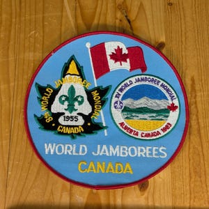 Może przedstawiać: Okrągła, niebieska naszywka z czerwonym obramowaniem, przedstawiająca tekst i grafikę związaną ze Światowymi Jamboree w Kanadzie. Zawiera flagę Kanady, kwiat lilii i napis "World Jamborees Canada". Naszywka ma średnicę około 10 cm.