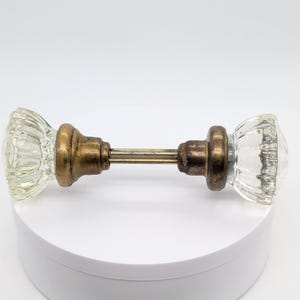 May include: A vintage door knob set featuring two clear, faceted glass knobs attached to a central metal spindle. Each knob is connected to a tarnished brass-coloured circular base plate, providing a classic hardware assembly for a door.