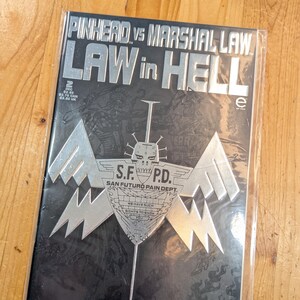 May include: A comic book titled "PINHEAD VS MARSHAL LAW LAW in HELL" with a black cover and silver lettering. The cover features a stylized emblem with the text "S.F.P.D. SAN FUTURO PAIN DEPT." and the words "WE HAVE SUCH SIGHTS TO SHOW YOU".
