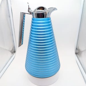 May include: A metallic blue thermal carafe with a ribbed, conical body and a shiny chrome-finished handle, lid, and spout. The base of the carafe is a solid white circular stand, providing a contrast to the vibrant blue textured exterior.