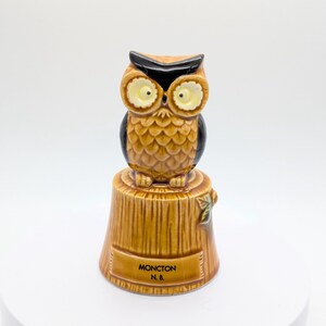 May include: A ceramic bell featuring a brown owl perched on a tree stump. The owl has large yellow eyes and black eyebrows. The base of the stump is textured like wood grain and displays the black text MONCTON N. B. on a rectangular plaque.