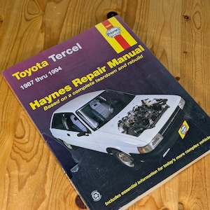 May include: A Haynes Repair Manual for the Toyota Tercel, covering the years 1987 through 1994. The cover features a white car with the hood open, showcasing the engine. The manual includes essential information for more complex vehicles.