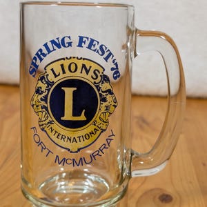 May include: A clear glass beer mug with a blue and gold logo that says "Spring Fest '76 Lions International Fort McMurray".