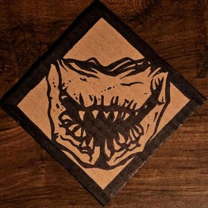 May include: A square, brown cardboard patch with a black border. The patch features a black and white illustration of a monster's open mouth with sharp teeth. The design is centered on the patch.