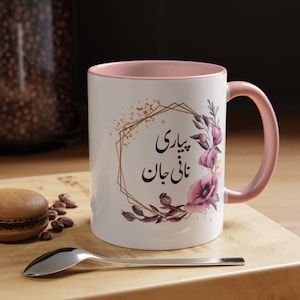 May include: White ceramic mug with a pink interior and handle. The mug features a floral design with the text "پياری نانی جان" in black script, framed by a gold geometric shape. A spoon and a macaron are in the foreground.
