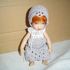 May include: A doll wearing a handmade gray crochet dress and bonnet. The doll has red hair and is posed sitting down. The crochet work features an open, lacy design. The doll is on a white surface with a brown background.