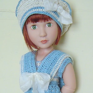 Puede incluir: Una muñeca vestida con un conjunto de ganchillo azul claro y blanco, que incluye un sombrero con un lazo blanco y un vestido a juego. La muñeca tiene el pelo rojo y ojos verdes, y un pequeño collar.