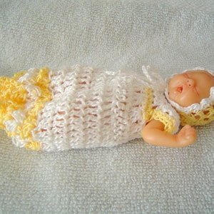 May include: A crocheted baby doll outfit in white and yellow with a matching crocheted hat. The doll is lying on a white towel.