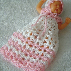 Puede incluir: Un vestido de muñeca de crochet con un patrón rosa y blanco. El vestido tiene un gorro de crochet a juego.