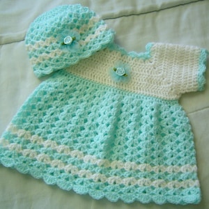 Puede incluir: Un conjunto de vestido y gorro de bebé tejido a crochet en azul claro y blanco con un adorno de flor.